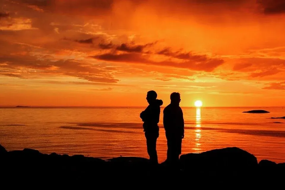 To personer i silhuett mot en oransje solnedgang ved sjøen